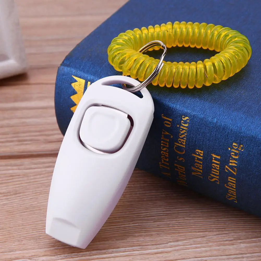 Clicker de dressage pour chiens de couleur blanc posé sur la tranche d'un livre bleu. Le produit est sur une table de couleur claire. Image produit Boutiquechien