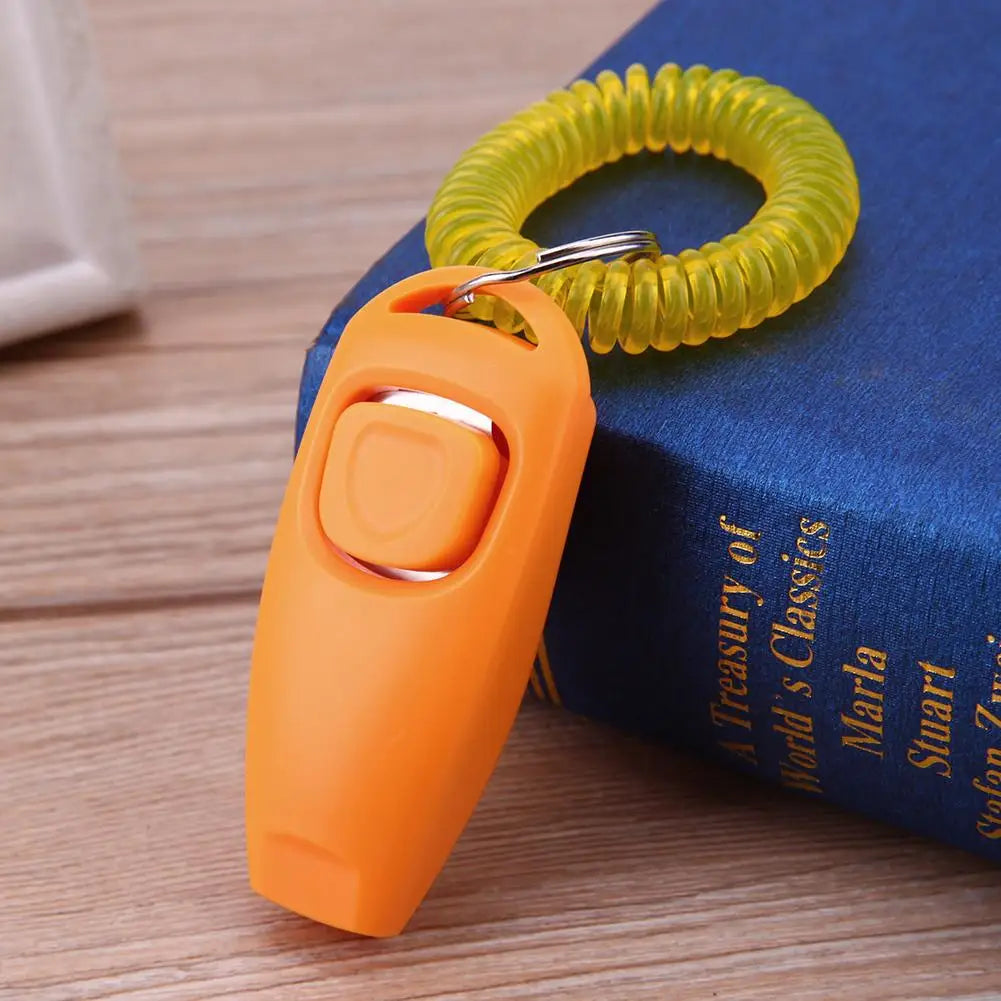 Clicker de dressage pour chiens de couleur orange posé sur un livre de couleur bleu. Le produit est sur une table de couleur claire. Image produit Boutiquechien