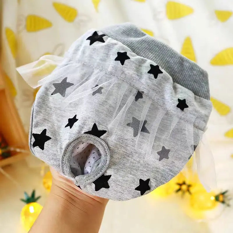 Couche pour chien de couleur grise avec des étoiles noir dessus tenu par une main. En fond il y a tissu blanc avec des carottes jaunes. Image produit Boutiquechien