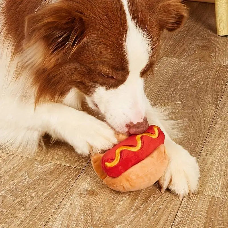 chien jouant avec sa peluche hot dog. Le chien est de couleur brun et blanc couché sur le sol en bois. Image produit Boutiquechien