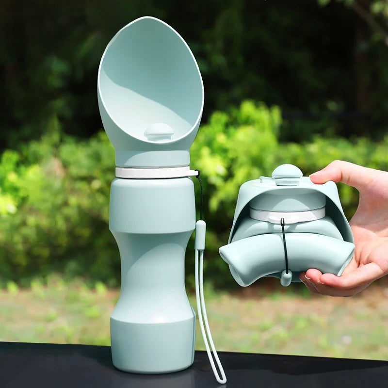 gourde pour chien de couleur verte pliante pour un rangement plus efficace. le produit est posé sur une table de couleur noir et maintenu par une main. En fond il y a une haie verte. Image produit Boutiquechien