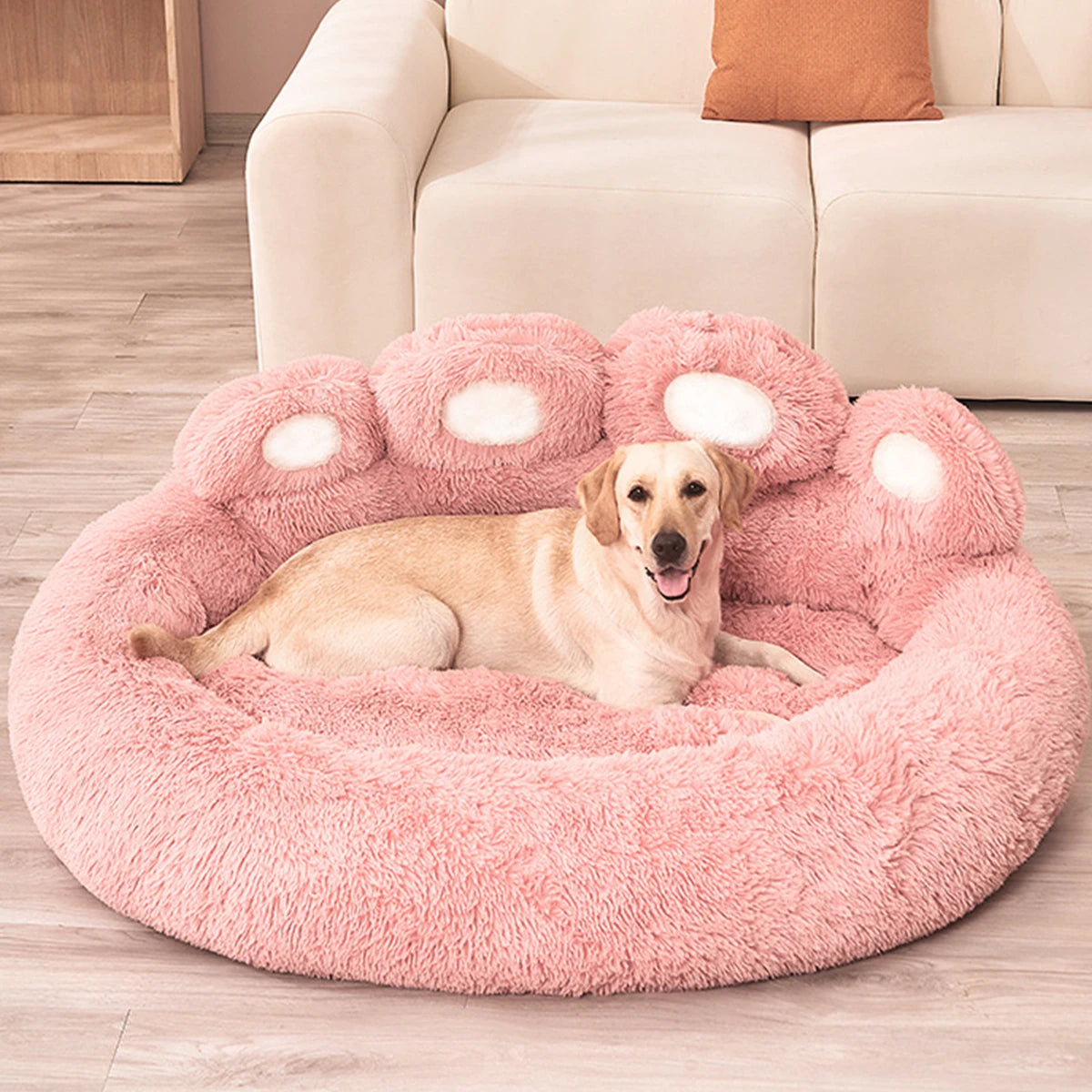 Gros chien couché dans le panier chien en forme de patte de couleur rose et blanc. Le panier chien est posé dans un salon avec en fond une canapé blanc. Image produit Boutiquechien