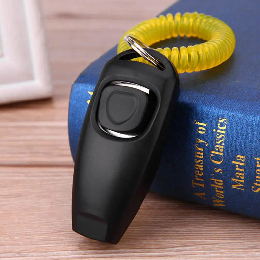 Clicker de dressage pour chiens de couleur noir posé sur la tranche d'un livre bleu. Le produit est sur une table de couleur claire. Image produit Boutiquechien