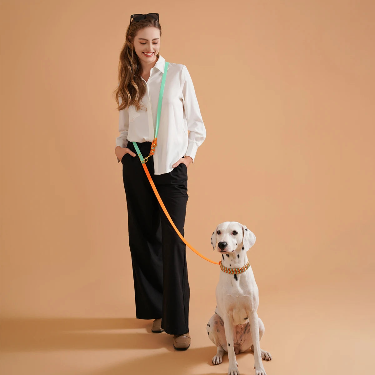 Femme souriante et élégante tenant la laisse en pvc résistante et durable. La laisse est de couleurs verte et orange  avec une femme habillé en noir et blanc. La laisse maintient un Dalmatien élégant assis. Image produit Boutiquechien