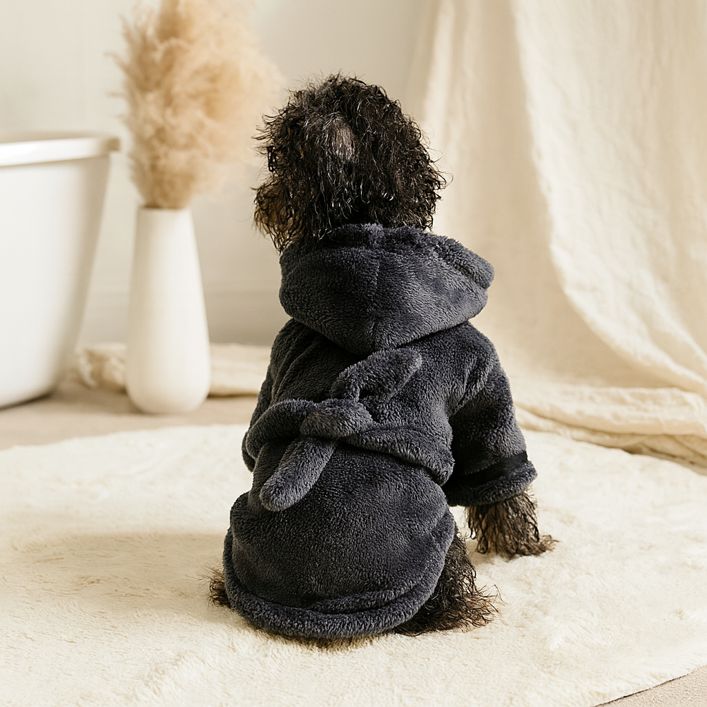 Petit chien noir mouillé portant un peignoir pour chien de couleur noir. Le chien est assis sur un tapis de bain de couleur blanc. avec ne fond une baignoire et une plante. Image produit Boutiquechien