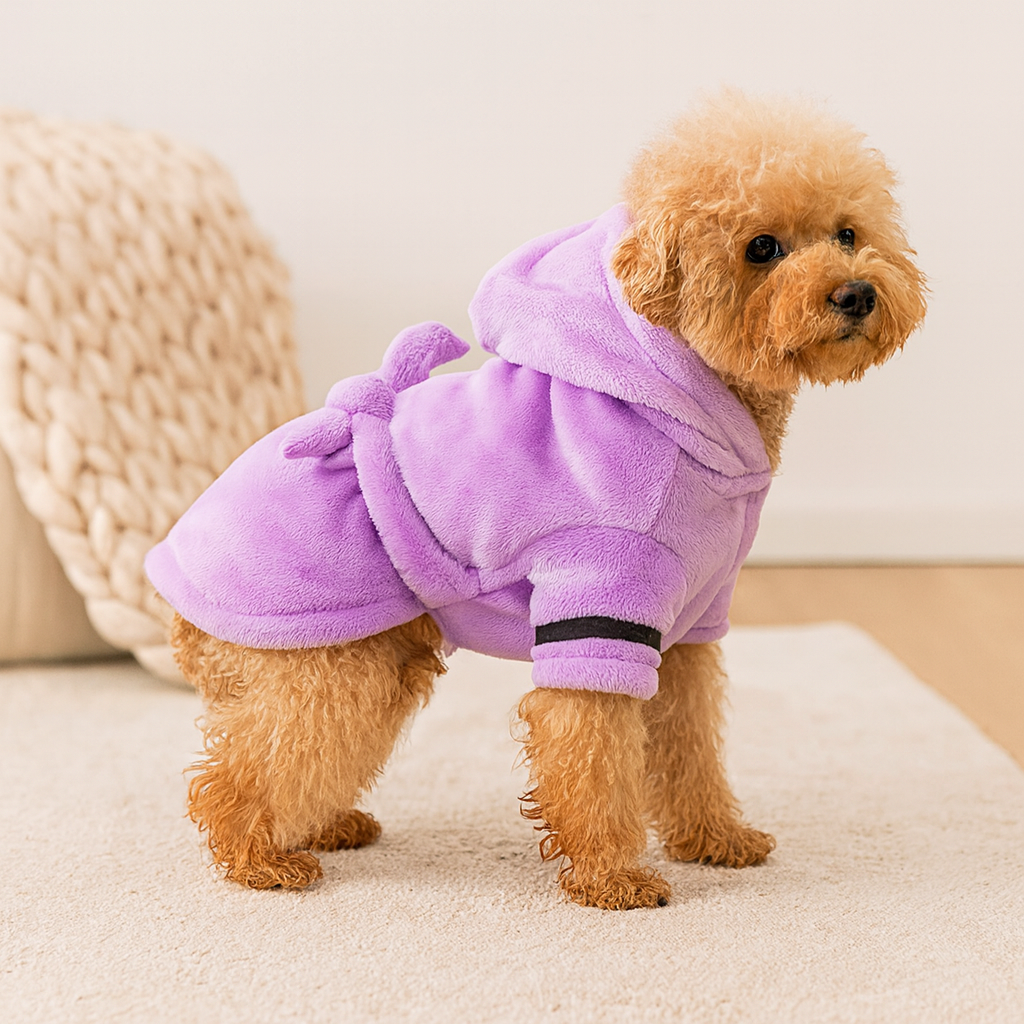 Petit chien de couleur claire avec un peignoir pour chien de couleur violet. Le chien est debout sur un tapis blanc avec en fond une laine blanche. Image produit Boutiquechien Image